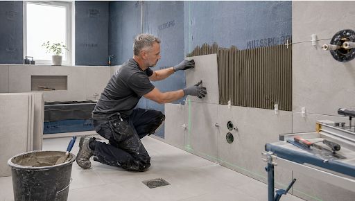 Construction worker kneels to tile a bathroom wall, placing tiles with spacers and using a trowel; bucket and tools nearby.