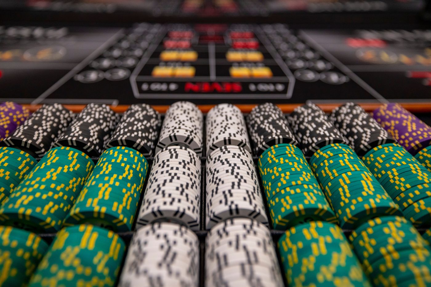 Rows of colorful poker chips (green, white, black) lined up on a casino table with a blurred betting layout in the background.