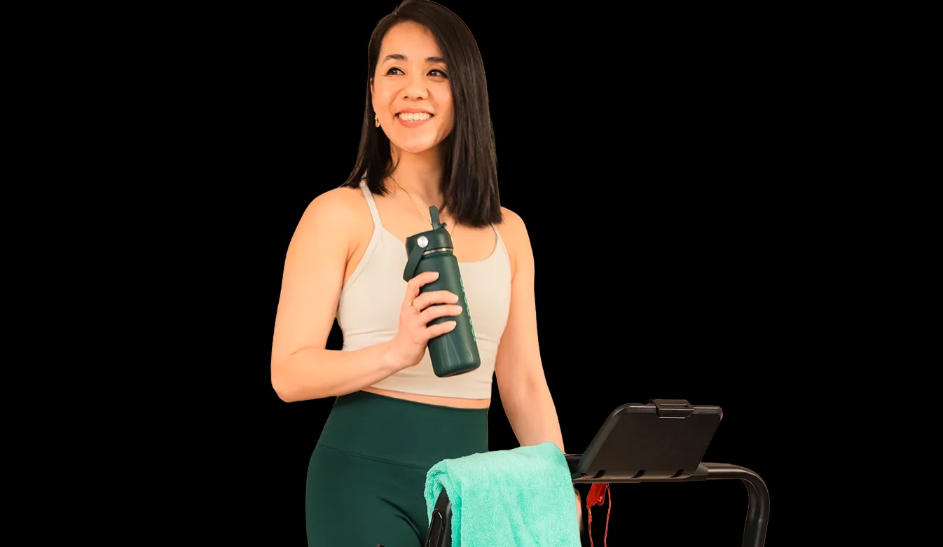 Smiling woman in a beige sports bra and green leggings holds a water bottle beside a treadmill with a mint towel draped over it.