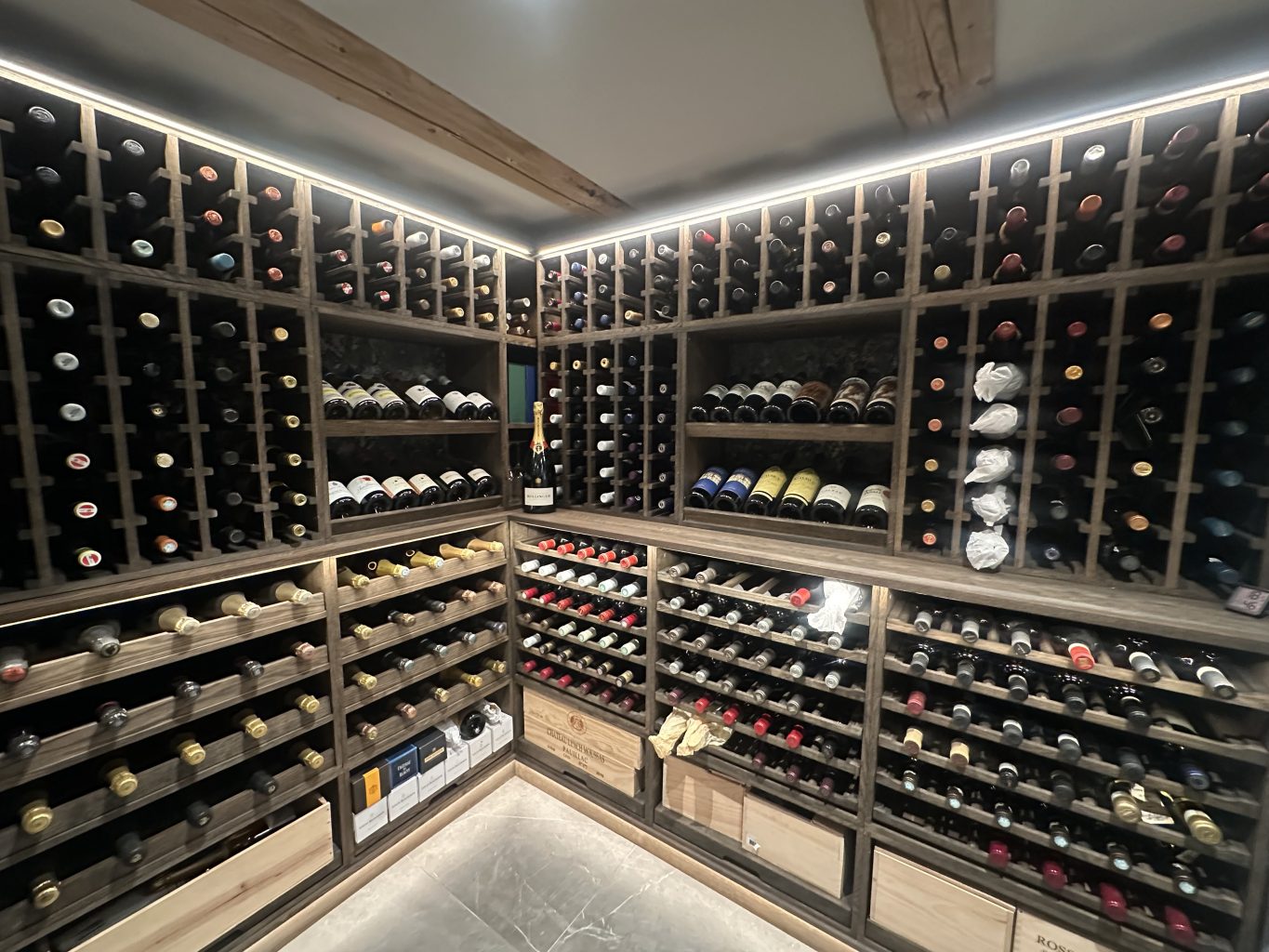 Corner wine cellar with wooden racks full of bottles arranged horizontally on all walls, a single champagne bottle standing on a middle shelf as a focal point.