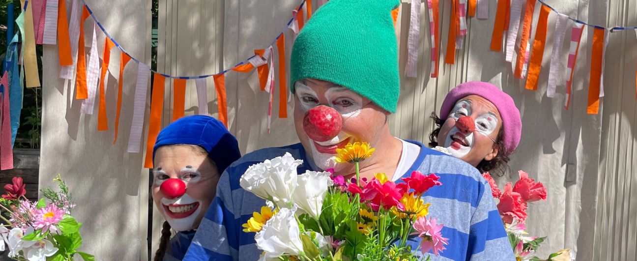Three clowns with painted faces and red noses pose with a bouquet of flowers, standing in front of orange-and-white streamers.
