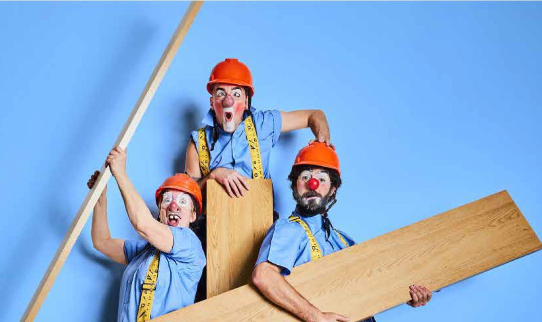 Three clowns in blue shirts and orange hard hats pose with wooden planks against a blue background, with measuring tapes around their necks for a humorous construction theme.