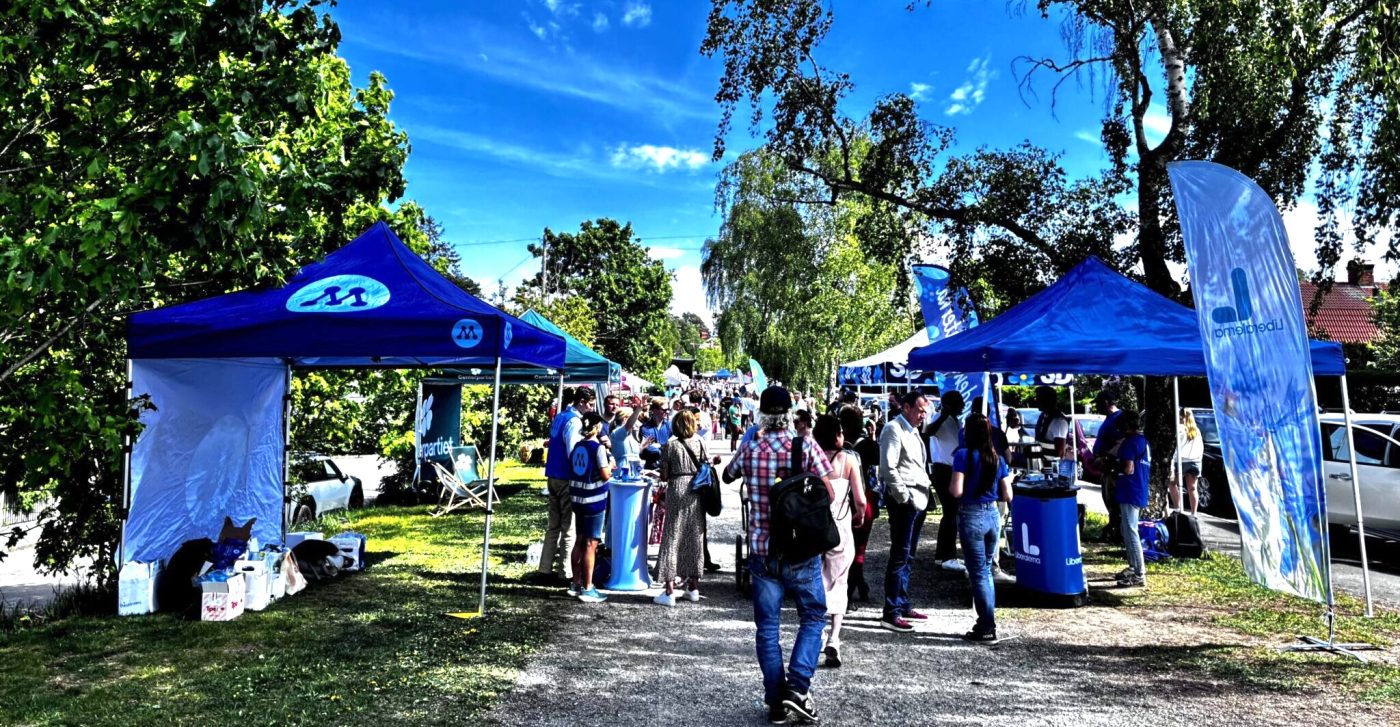 Outdoor street fair with blue canopies and attendees visiting booths on a sunny day.