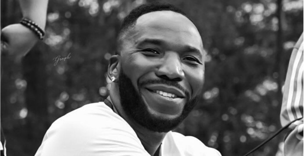 Close-up portrait of a smiling man with a beard outdoors, wearing a white shirt and an earring in one ear.