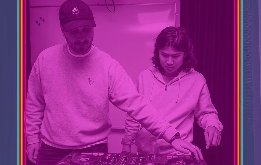 Two people wearing hoodies operate a DJ mixer and turntables in a purple-lit roomが。