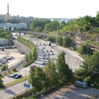 Aerial view of a multi-lane highway with cars, beside a railway overpass and green, tree-lined hills in a suburban cityscape.