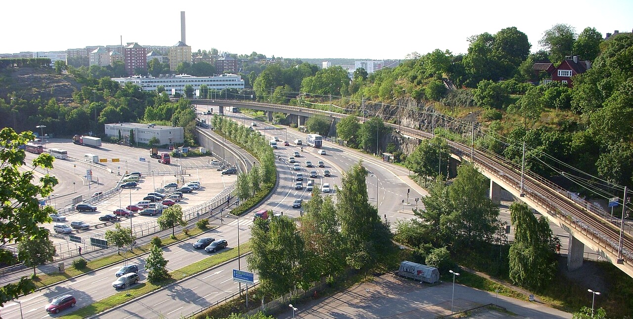 Aerial view of a multi-lane highway with cars, beside a railway overpass and green, tree-lined hills in a suburban cityscape.