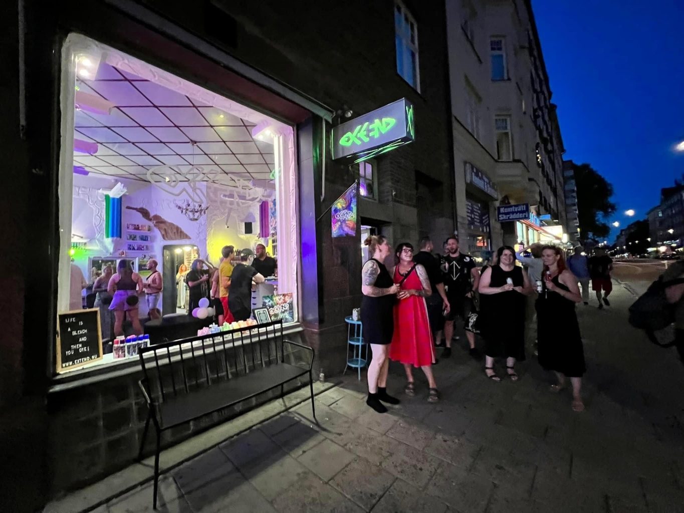 Night scene: people mingle outside a neon-lit storefront bar on a city street at dusk; colorful interior visible through the window.