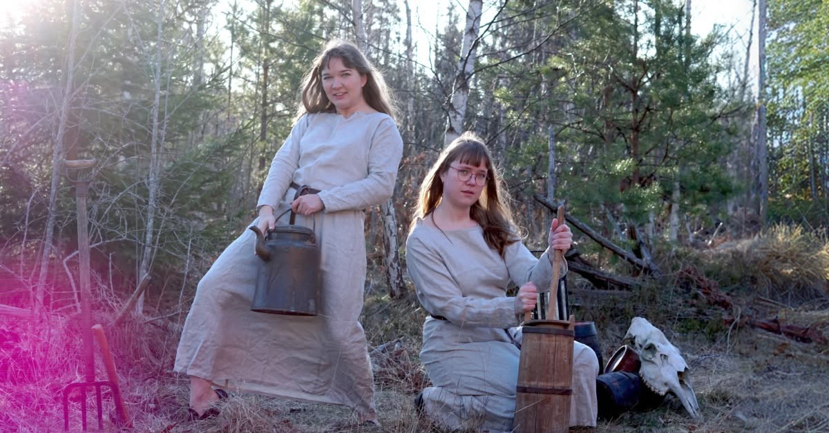 Two women in beige linen dresses stand and sit in a sunlit forest, handling vintage milking equipment (metal kettle and wooden churn).