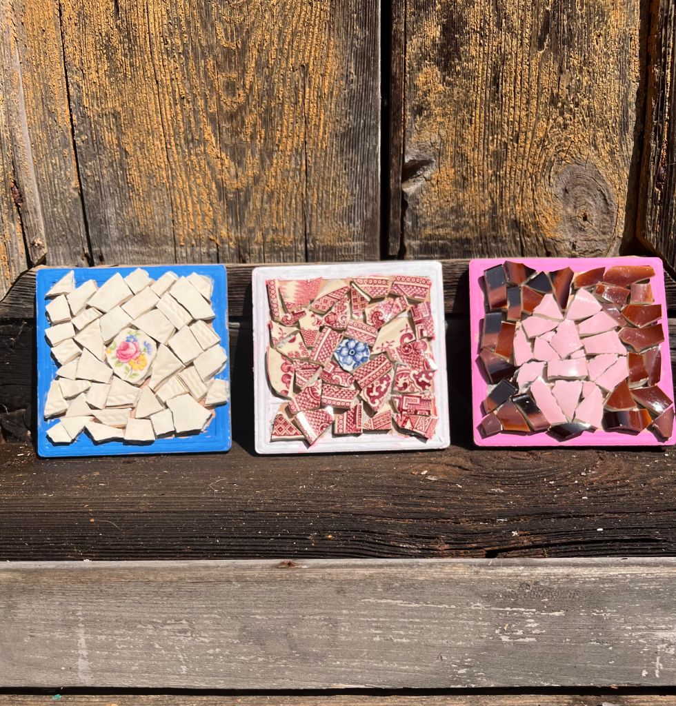 Three trays of mosaic tiles on a rustic wooden surface: blue tray with beige squares, white tray with red patterned pieces, pink tray with pink and brown shards.