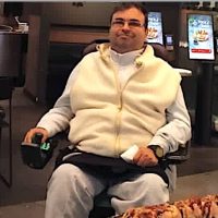 Man in a wheelchair sits at a restaurant table with a tray of sushi in front of him, wearing a white zip‑up vest and light sweatpants.