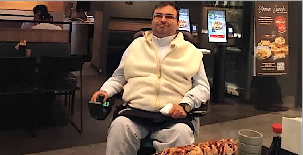 Man in a wheelchair sits at a restaurant table with a tray of sushi in front of him, wearing a white zip‑up vest and light sweatpants.
