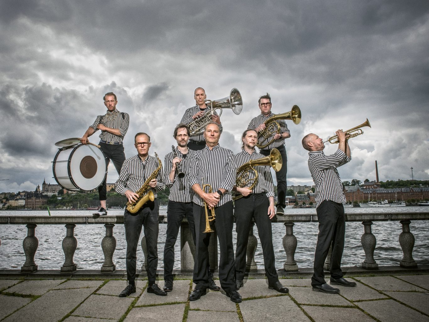 Seven musicians in striped shirts pose with brass instruments on a riverside promenade under a cloudy sky.