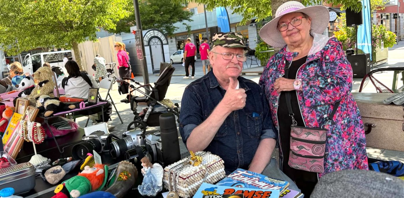 Two elderly people at an outdoor market stall with toys, cameras, and trinkets; man gives a thumbs‑up while seated beside a woman in a wide hat and floral jacket.