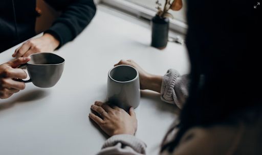 Two people share a moment at a white table, passing gray mugs as they chat, with a small plant in the background.