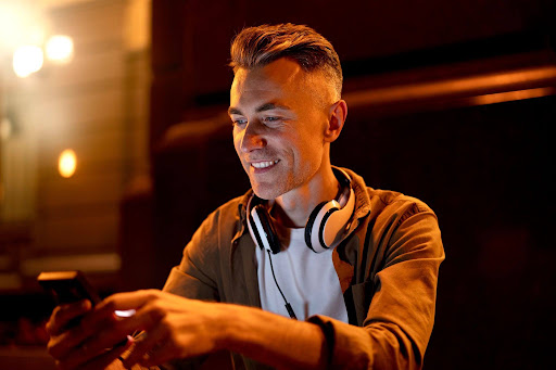 Man wearing headphones around his neck smiles while looking at a smartphone in a warm, dimly lit room.